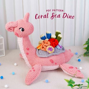 Könnte beinhalten: Ein handgefertigtes, rosa gehäkeltes Dinosaurier-Spielzeug mit einem Korallenriff-Design auf dem Rücken. Der Dinosaurier hat einen langen Hals und einen Schwanz. Der Text "PDF PATTERN Coral Sea Dino" ist sichtbar.