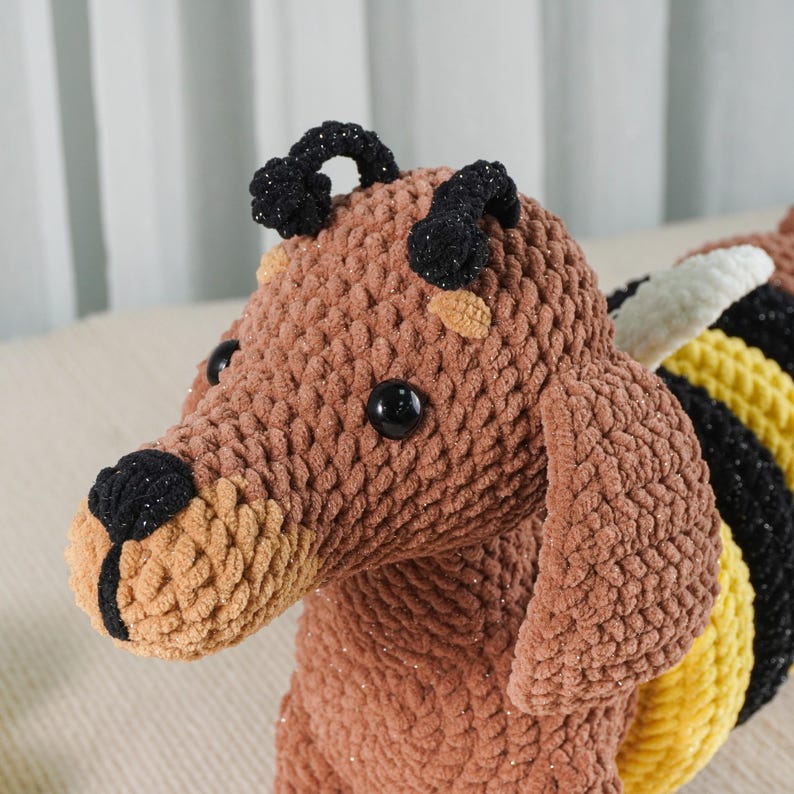 May include: A handmade brown crocheted bee-dog plush toy. The toy features black eyes, a black nose, and black antennae. The bee-dog has yellow, black, and white striped wings. The plush toy is made of soft, textured yarn.