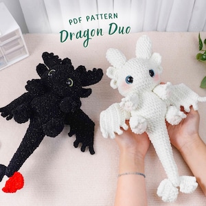 May include: Two crocheted dragon toys, one black and one white, are displayed. The black dragon has a red tail accent. The white dragon is held in a person's hands. The text "PDF PATTERN Dragon Duo" is at the top.