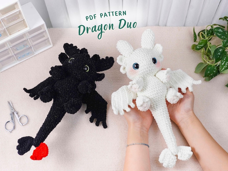 May include: Two crocheted dragon toys, one black and one white, are displayed. The black dragon has a red tail accent. The white dragon is held in a person's hands. The text "PDF PATTERN Dragon Duo" is at the top.