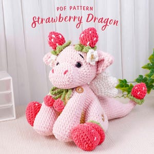 May include: A pink crocheted dragon with strawberry details. The dragon has white wings, a green scarf, and red strawberry accents on its head, feet, and tail. The text "PDF PATTERN Strawberry Dragon" is displayed above the dragon.