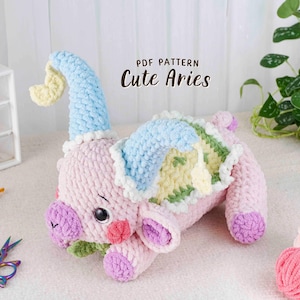 May include: A handmade, plush Aries toy in soft pink, blue, and yellow. The crochet animal has a light blue hat, a green and yellow back, and purple accents. The text "PDF Pattern Cute Aries" is displayed on the image.