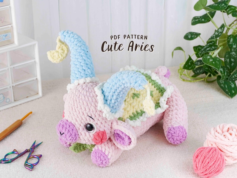 May include: A handmade, plush Aries toy in soft pink, blue, and yellow. The crochet animal has a light blue hat, a green and yellow back, and purple accents. The text "PDF Pattern Cute Aries" is displayed on the image.