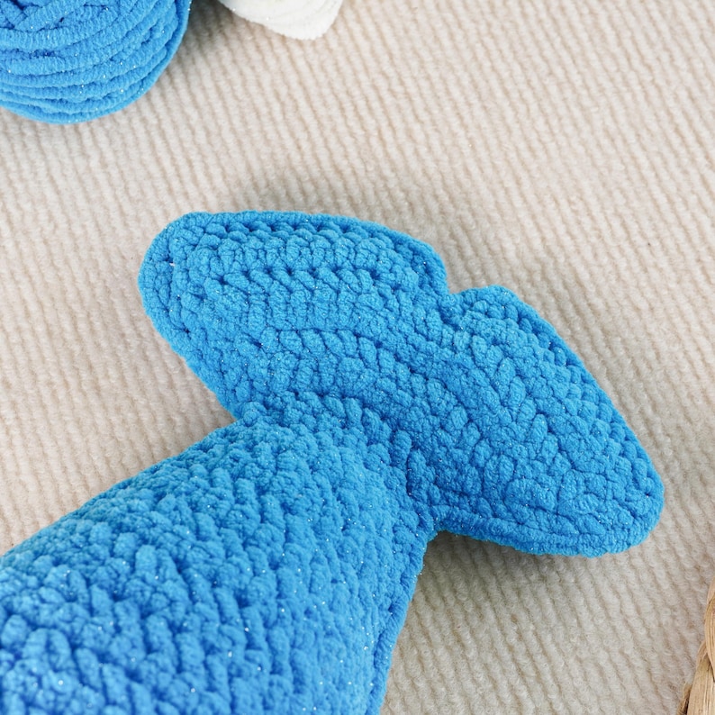 May include: Close-up of a bright blue, crocheted object with a tail-like shape. The object is made of a soft, textured yarn. The background is a light beige, textured surface. Other crocheted items are visible.