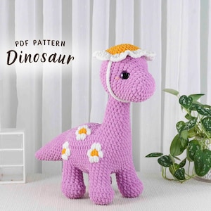 May include: A handmade, lavender-colored crochet dinosaur with white and yellow flower accents. The dinosaur wears a yellow and white hat. The image includes the text "PDF PATTERN Dinosaur".
