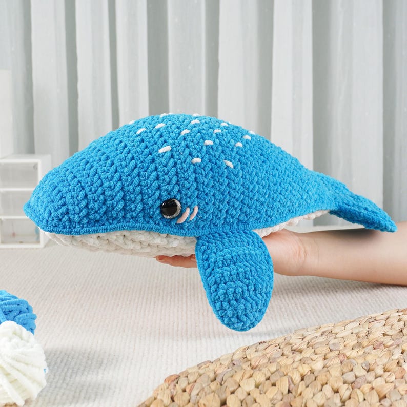 May include: A handmade blue crochet whale plush toy with white accents. The whale features a textured body, white belly, and small white spots on its back. It has a small eye and a fin on each side. A soft, cuddly ocean-themed toy.