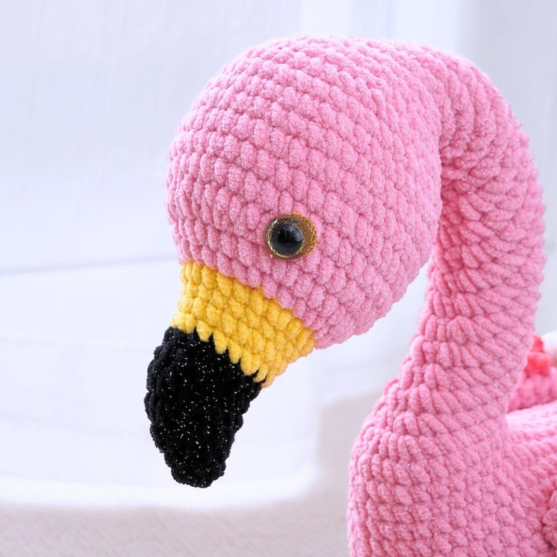 May include: A handmade pink crochet flamingo with a yellow and black beak. The flamingo has a large, dark eye and is made of soft, textured yarn. The flamingo is a whimsical and decorative item.