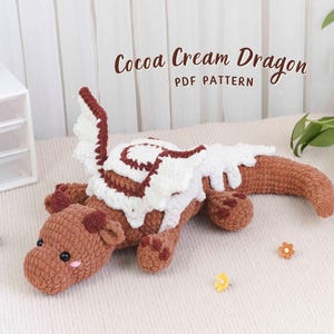 Może przedstawiać: Szydełkowa zabawka smoka w odcieniach brązu, bieli i czerwieni. Smok ma fantazyjny design z kremowym brzuchem i skrzydłami. Tekst "Cocoa Cream Dragon PDF Pattern" jest wyświetlany nad smokiem.