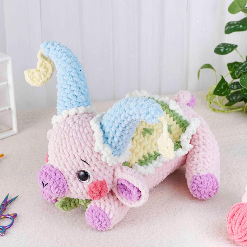 May include: A handmade pink crocheted piglet with a blue and yellow hat and a colorful blanket. The piglet has purple hooves, a green leaf, and a white trim. The toy is lying down on a white surface.