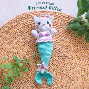 May include: A white crocheted mermaid kitten with a pink flower on its head and a turquoise tail. The kitten is wearing a pink and purple bikini top. The kitten is sitting on a woven straw mat.