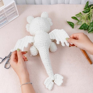 May include: A white crocheted dragon toy with outstretched wings, tail, and small feet. The dragon is made of a soft, textured yarn. The wings are being attached with scissors. The background is a light pink surface.