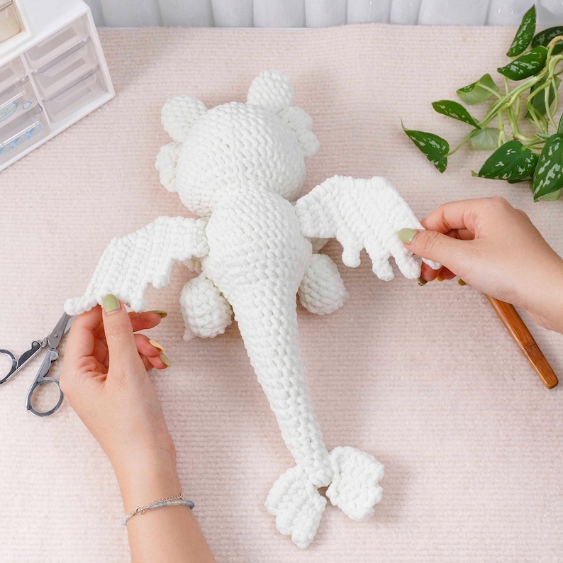 May include: A white crocheted dragon toy with outstretched wings, tail, and small feet. The dragon is made of a soft, textured yarn. The wings are being attached with scissors. The background is a light pink surface.