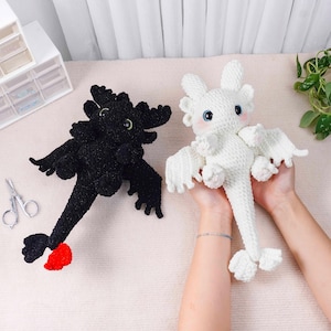 May include: Two crocheted dragon plushies, one black and one white, are displayed. The black dragon has a red tail fin and green eyes. The white dragon has blue eyes and pink cheeks. Both have wings and are handmade.