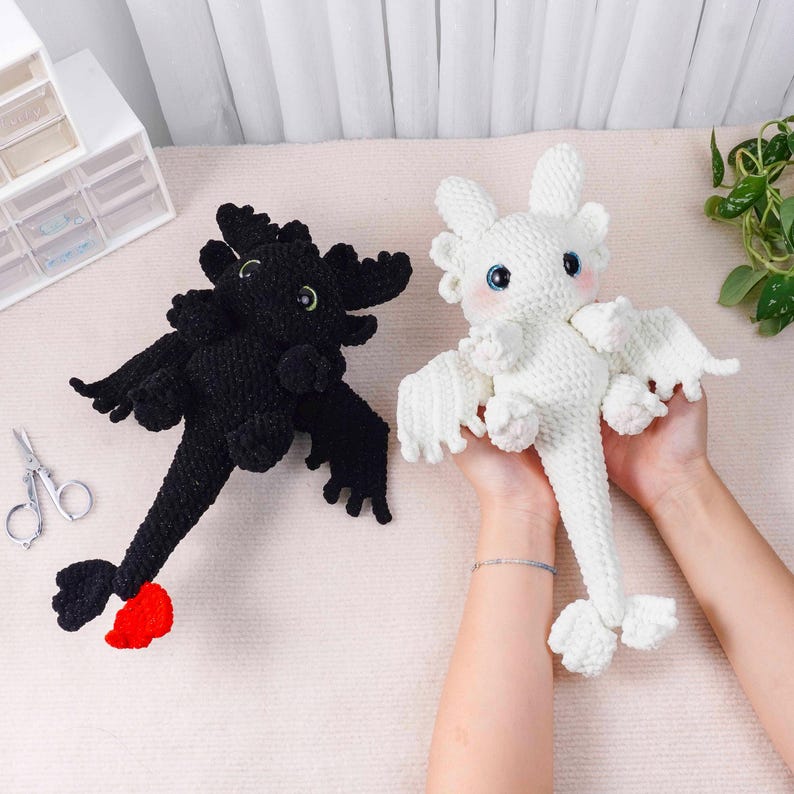 May include: Two crocheted dragon plushies, one black and one white, are displayed. The black dragon has a red tail fin and green eyes. The white dragon has blue eyes and pink cheeks. Both have wings and are handmade.