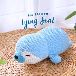 Può includere: Un peluche di foca all'uncinetto blu chiaro in posizione sdraiata. La foca ha pancia e viso bianchi con occhi neri e naso nero. Il testo "PDF PATTERN Lying Seal" è sopra la foca.