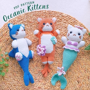 Peut inclure: Trois chatons en crochet rembourrés, un bleu et blanc, un orange et blanc, et un blanc avec une queue turquoise et un haut rose et violet. Les chatons sont sur un tapis de paille tissé. Le texte "Oceanic Kittens PDF Pattern" est au-dessus des chatons.
