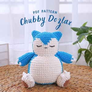 Pode incluir: Padrão PDF para um brinquedo de pelúcia Snorlax de crochê azul e branco. O brinquedo está sentado em um tapete de palha trançado.