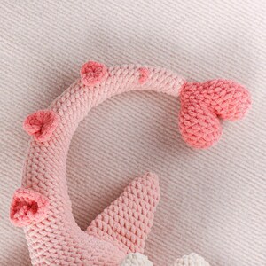 May include: A close-up of a pink crocheted plush toy, possibly a sea creature or a whimsical design. The toy features a curved body with three round, textured accents and a tail-like appendage. The plush is made of soft, textured yarn.