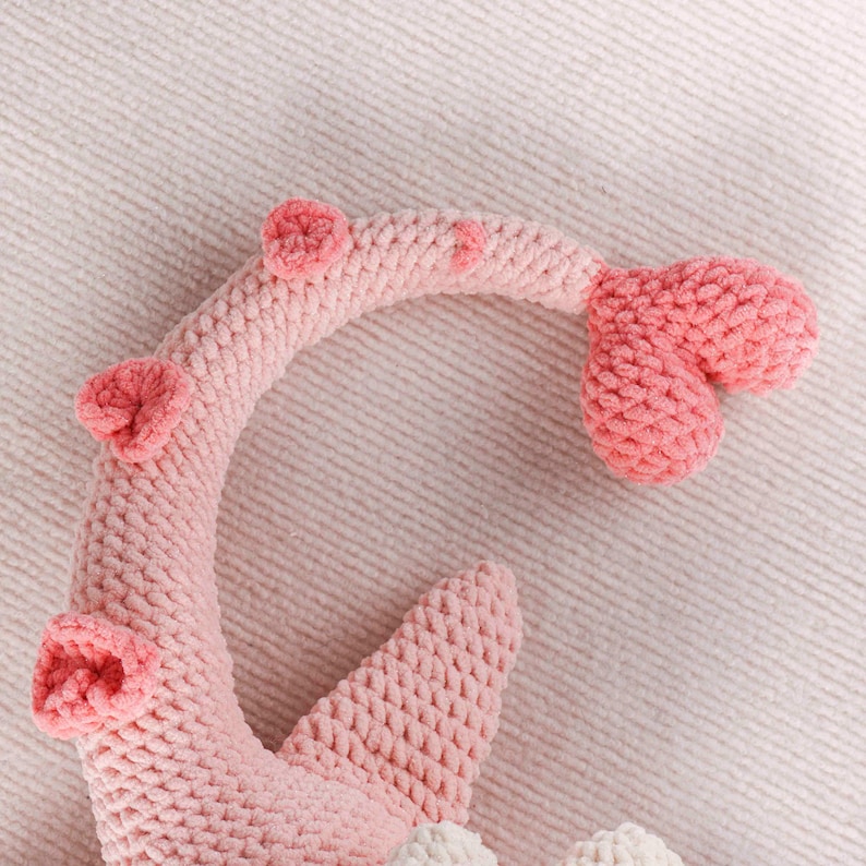 May include: A close-up of a pink crocheted plush toy, possibly a sea creature or a whimsical design. The toy features a curved body with three round, textured accents and a tail-like appendage. The plush is made of soft, textured yarn.