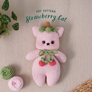 May include: A pink crocheted strawberry cat amigurumi toy with green leaves on its head and a green bow. The toy is sitting on a beige surface. PDF PATTERN strawberry Cat