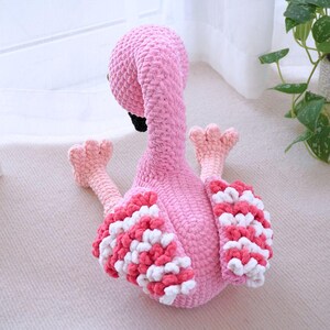 May include: A handmade pink flamingo plush toy with a textured body, black beak, and pink and white wing accents. The flamingo is sitting down with its legs extended, showcasing its detailed crochet work.