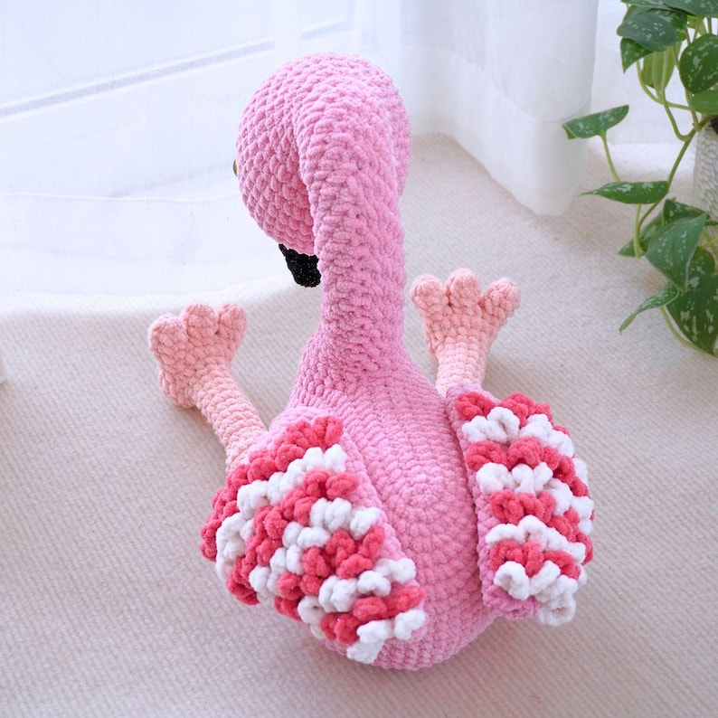 May include: A handmade pink flamingo plush toy with a textured body, black beak, and pink and white wing accents. The flamingo is sitting down with its legs extended, showcasing its detailed crochet work.