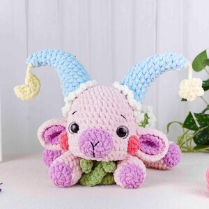 May include: A handmade crochet goat plushie in soft pink, purple, and blue. The plushie has large, round eyes, a purple nose, and a green leafy collar. It wears blue horns with yellow accents, and is a whimsical toy.