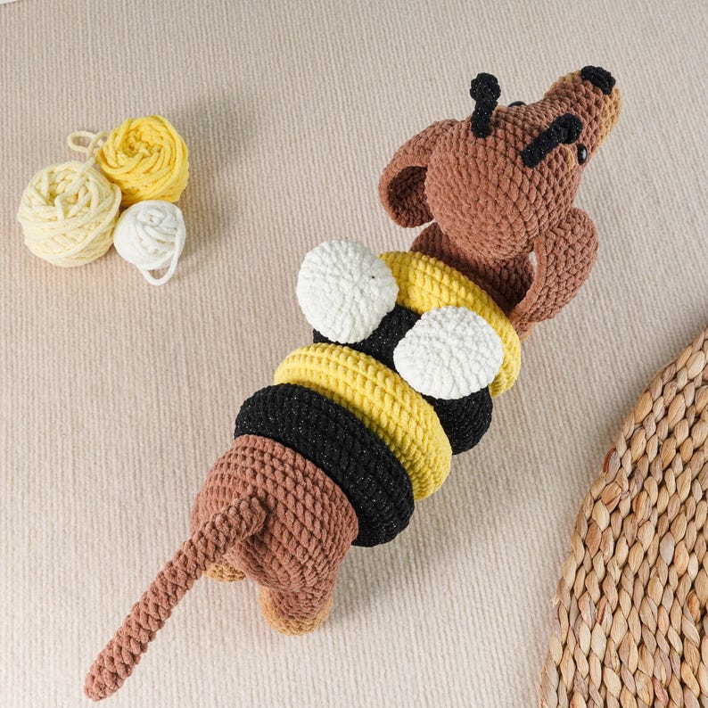 May include: A handmade crochet giraffe toy in shades of brown, yellow, black, and white. The giraffe has a long neck and tail, with a series of colorful rings forming its body. Yarn balls in yellow and white are in the background. A whimsical and playful design.