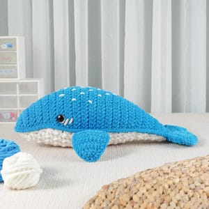 May include: A handmade blue and white crochet whale plush toy. The whale features a blue body with white spots and a white belly. It has small black eyes and a smiling mouth. The toy is made of soft yarn and is designed to be a cuddly item.
