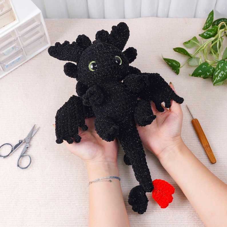 May include: A handmade black crochet dragon plush toy with green eyes and a red tail fin. The dragon has large wings and a detailed head with multiple horns. The plush is held in someone's hands, with a pair of scissors and a crochet hook visible.