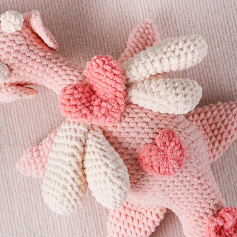 May include: A soft, pink crochet unicorn toy with white details and heart shapes. The handmade toy has a fluffy texture and is designed with a whimsical aesthetic, suitable for a child's room or as a decorative item.