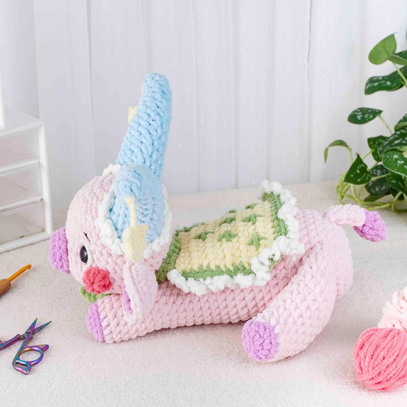 May include: A handmade pink crochet elephant toy with blue, green, and yellow accents. The elephant has a trunk, ears, and a decorative blanket on its back. The toy is lying down on a white surface.