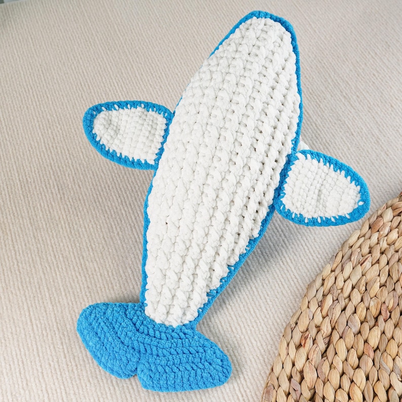 May include: A handmade, crocheted airplane toy in white and blue. The body is white, outlined in blue, with blue wings and tail. The soft toy is designed to resemble a classic airplane.