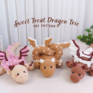 May include: Three crocheted dragon toys in shades of pink, brown, and tan, with white accents. The dragons have wings and are lying down. The text "Sweet Treat Dragon Trio PDF Pattern" is displayed above the toys.