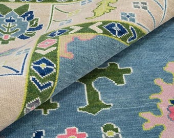 Könnte beinhalten: Nahaufnahme eines aufgerollten Teppichs mit detailliertem Muster. Der Teppich hat einen hellbeigen Hintergrund mit blauen, grünen und rosa floralen und geometrischen Designs. Der gerollte Bereich zeigt die Textur und Farbvariationen des Teppichs.