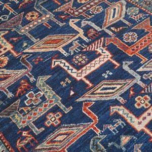 May include: Close-up of a blue rug featuring a repeating pattern of stylized birds and geometric shapes in red, beige, and grey. The rug has a border with floral and geometric designs.