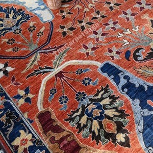 May include: An intricately designed rug with a vibrant color palette. The rug features a terracotta background with detailed floral and botanical patterns in shades of blue, brown, and cream. A decorative vase sits atop the rug.