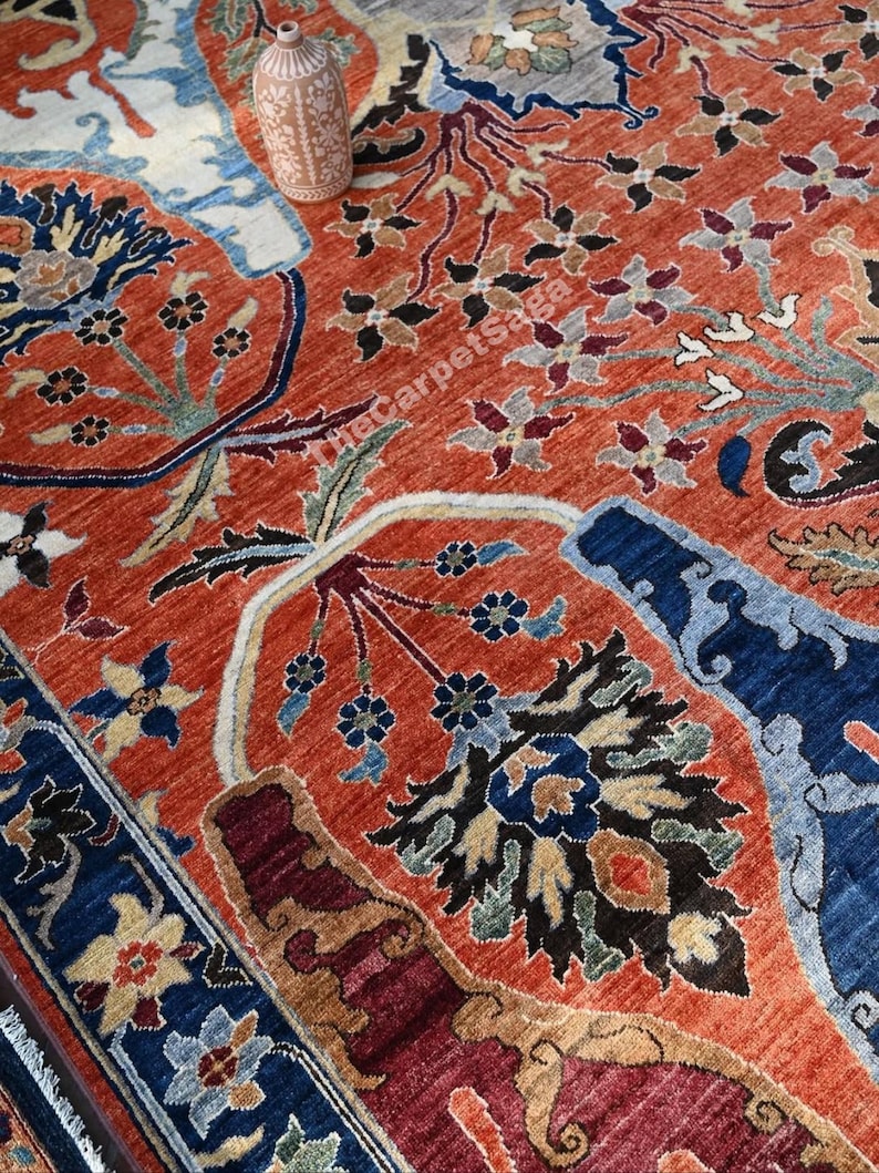 May include: An intricately designed rug with a vibrant color palette. The rug features a terracotta background with detailed floral and botanical patterns in shades of blue, brown, and cream. A decorative vase sits atop the rug.