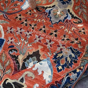 May include: A detailed shot of a hand-knotted rug with a vibrant, warm color palette. The rug features intricate floral and geometric patterns in shades of red, blue, black, and cream. A small, decorative vase sits atop the rug.