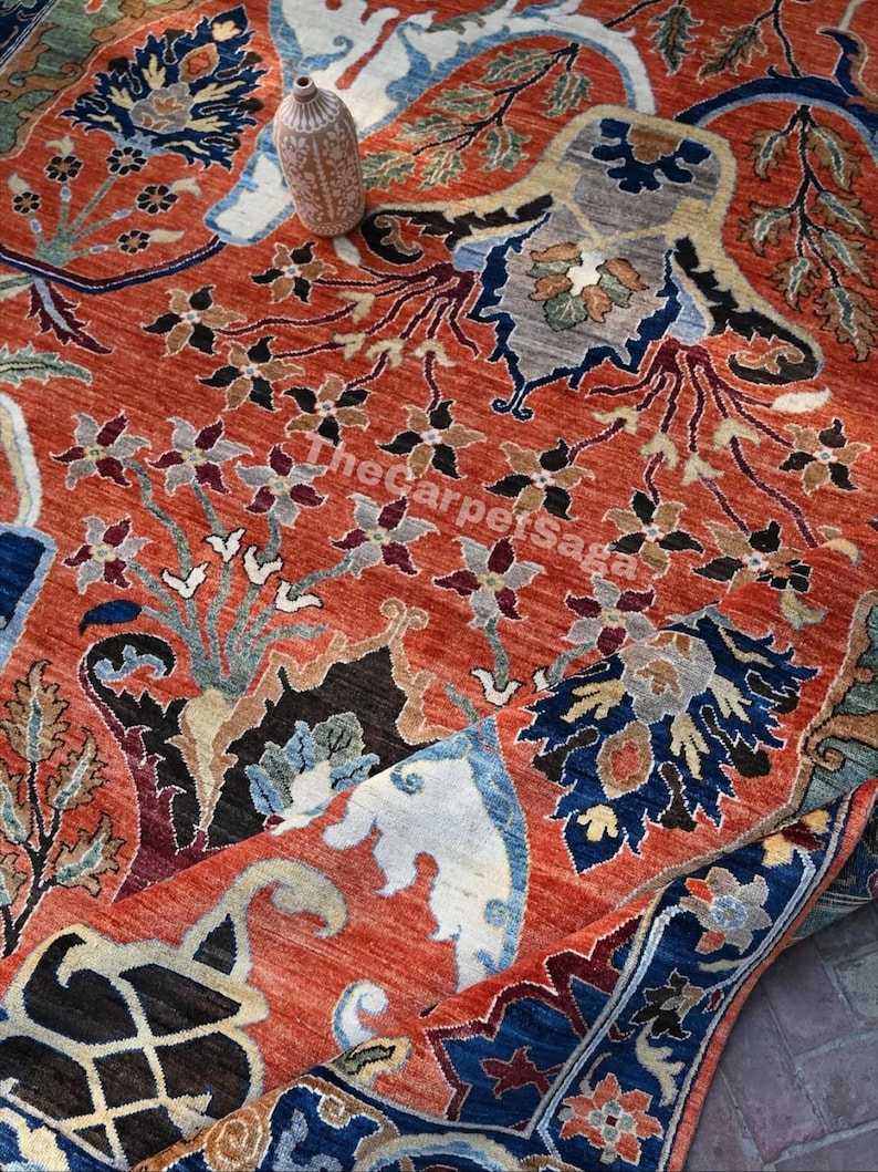 May include: A detailed shot of a hand-knotted rug with a vibrant, warm color palette. The rug features intricate floral and geometric patterns in shades of red, blue, black, and cream. A small, decorative vase sits atop the rug.