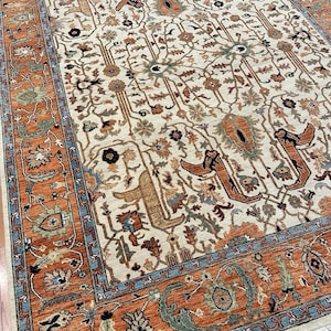 May include: An intricately patterned rug with a cream-colored field and a border in shades of orange, blue, and green. The rug features stylized floral and geometric motifs, creating a traditional aesthetic. The rug's design includes repeating patterns and detailed elements.