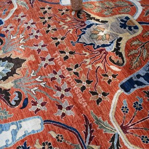 May include: An intricately patterned, rust-colored rug with floral and geometric designs in shades of blue, beige, and black. A decorative vase sits atop the rug. The text "The Carpersaga" is visible on the rug.
