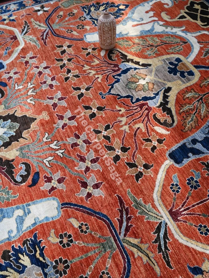 May include: An intricately patterned, rust-colored rug with floral and geometric designs in shades of blue, beige, and black. A decorative vase sits atop the rug. The text "The Carpersaga" is visible on the rug.