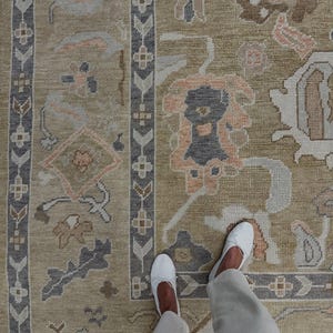 May include: A beige area rug with a detailed floral pattern in shades of gray, peach, and brown. The rug has a border with a geometric design. White shoes and light-colored pants are visible.