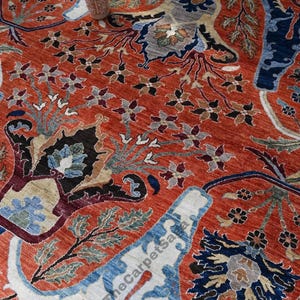 May include: Close-up of a vibrant, intricately patterned rug. The rug features a rich terracotta background with detailed floral and foliate designs in shades of blue, cream, and black. A decorative vase sits atop the rug.