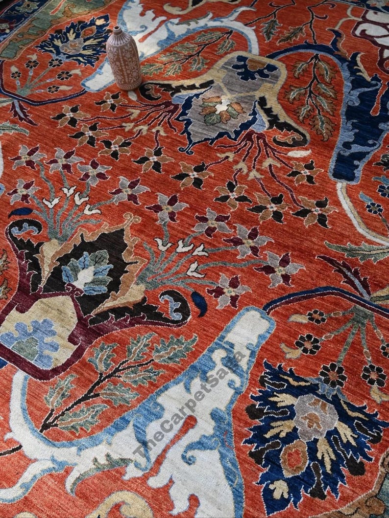 May include: Close-up of a vibrant, intricately patterned rug. The rug features a rich terracotta background with detailed floral and foliate designs in shades of blue, cream, and black. A decorative vase sits atop the rug.