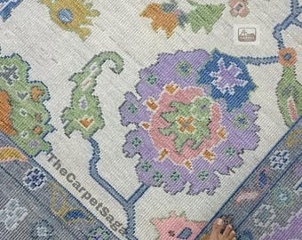 May include: A white rug with a blue, green, pink, and orange floral pattern. The rug has a faded look and is worn in some areas. The rug is on a wooden floor and a person's bare feet are visible in the bottom right corner of the image.