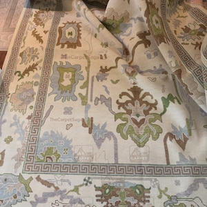 May include: A large area rug with a cream-colored background featuring a repeating Greek key border and stylized floral motifs in shades of sage green, light blue, and brown. The text TheCarpetSaga is visible in multiple locations on the rug.