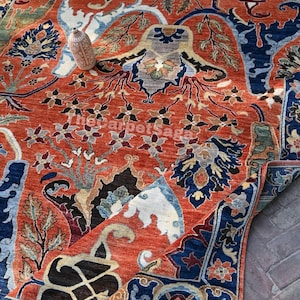 May include: An intricately designed area rug with a rich, warm color palette. The rug features a predominantly rust-colored field with detailed floral and geometric patterns in navy blue, cream, and touches of green. A small vase rests on the rug.