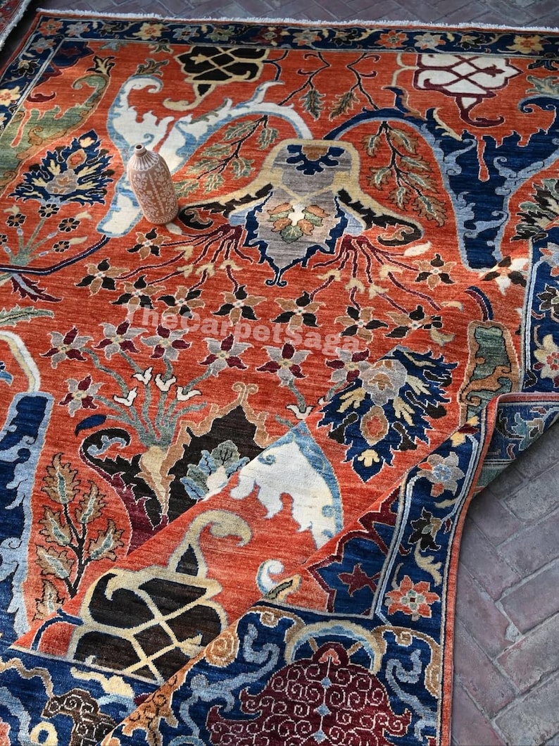 May include: An intricately designed area rug with a rich, warm color palette. The rug features a predominantly rust-colored field with detailed floral and geometric patterns in navy blue, cream, and touches of green. A small vase rests on the rug.
